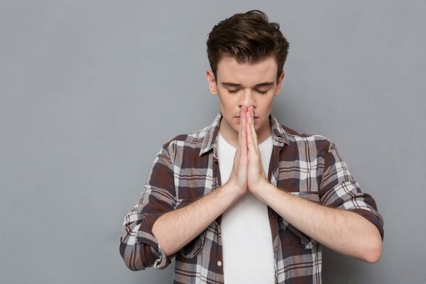 Concentrated stressed handsome young man in checkered shirt praying with closed eyes