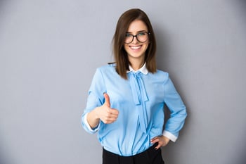 Happy businesswoman showing thumb up over gray background. Wearing in blue shirt and glasses. Looking at camera-1