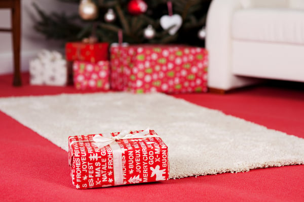 Red Christmas gift lying alone on a living room floor on the edge of a white rug overlaying a red carpet with additional gifts and the bottom of the decorated Christmas tree in the distance